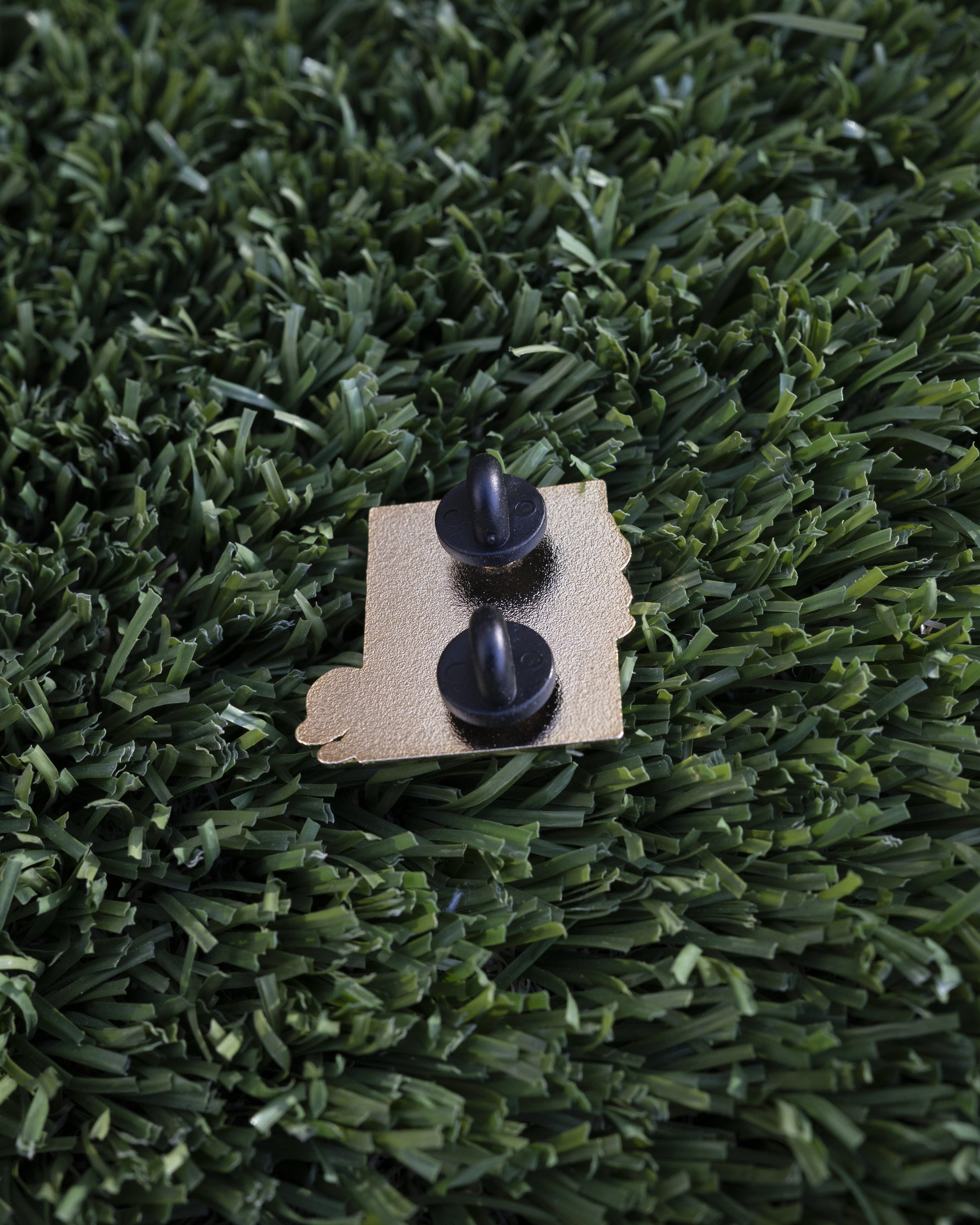 Hitorii "Spray-proof" metal chalkbag pin shown from the back on a backdrop of artrificial grass. The pin has a plain metal back with two pins, lined up vertically, for extra stability.