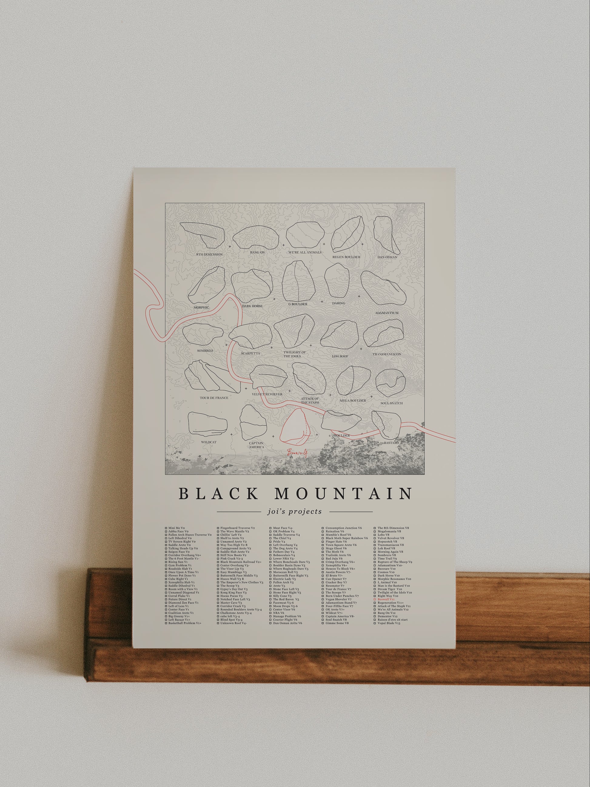 A cream color, customized rock climbing ticklist poster for the Black Mountain rock climbing location in San Jacinto Mountain, California. The design has an overlay of crag topography, nature landscapes, and traced boulders, the person's name, and a list of bouldering problems at the bottom. Displayed as wall art in a living room.