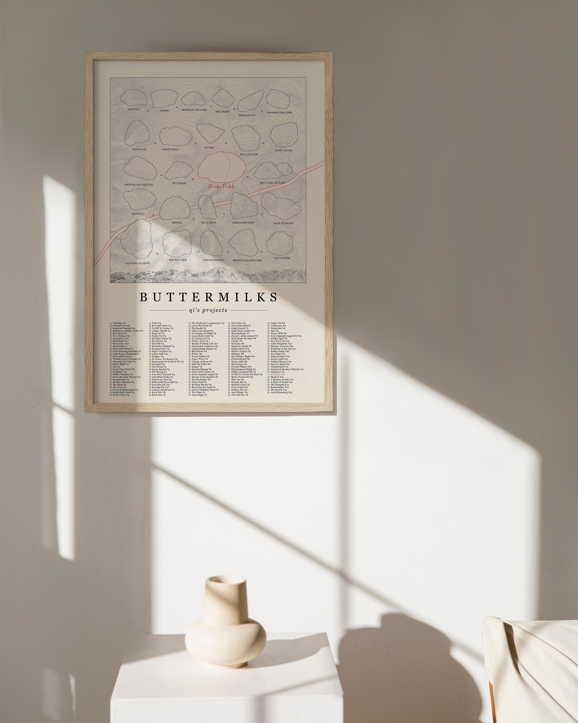 A cream color, customized rock climbing ticklist poster for the Buttermilks rock climbing location in Bishop, California. The design has an overlay of crag topography, nature landscapes, and traced boulders, the person's name, and a list of bouldering problems at the bottom. Displayed as wall art in a living room.