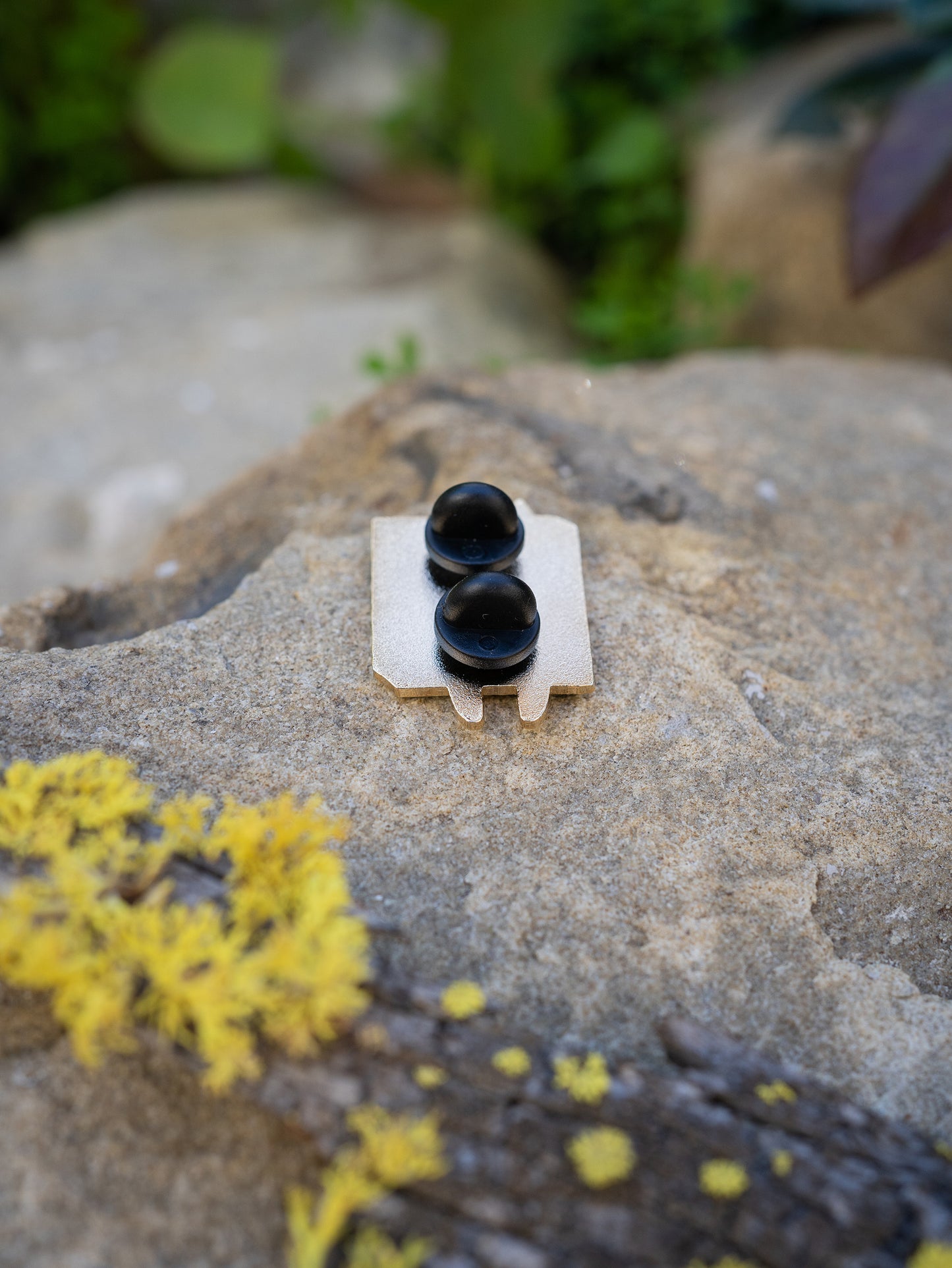 Hitorii "Crashpad Mao" metal chalkbag pin shown from the back on a backdrop of a rock. The pin has a plain metal back with two pins, lined up vertically, for extra stability.
