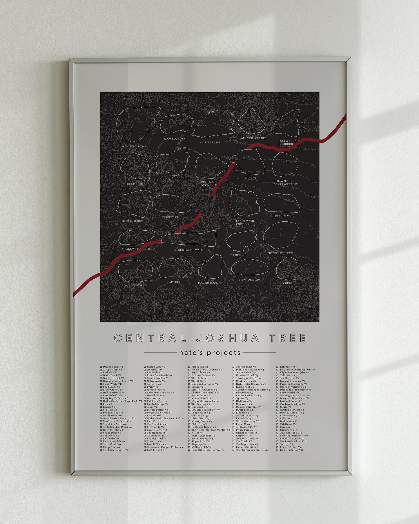 A black color, customized rock climbing ticklist poster for the JTree Central rock climbing location in Joshua Tree, California. The design has an overlay of crag topography, nature landscapes, and traced boulders, the person's name, and a list of bouldering problems at the bottom. Displayed as wall art in a living room.