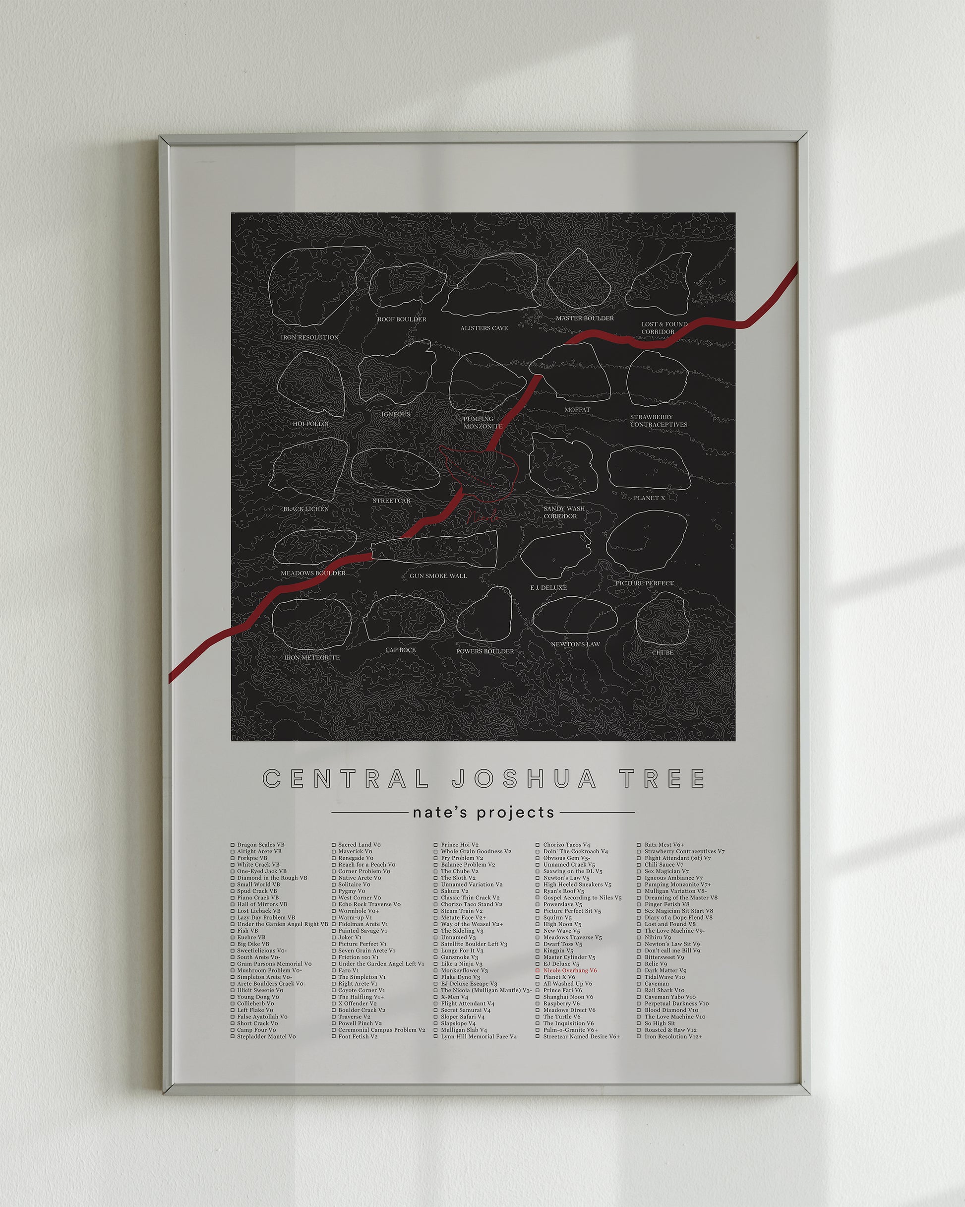 A black color, customized rock climbing ticklist poster for the JTree Central rock climbing location in Joshua Tree, California. The design has an overlay of crag topography, nature landscapes, and traced boulders, the person's name, and a list of bouldering problems at the bottom. Displayed as wall art in a living room.