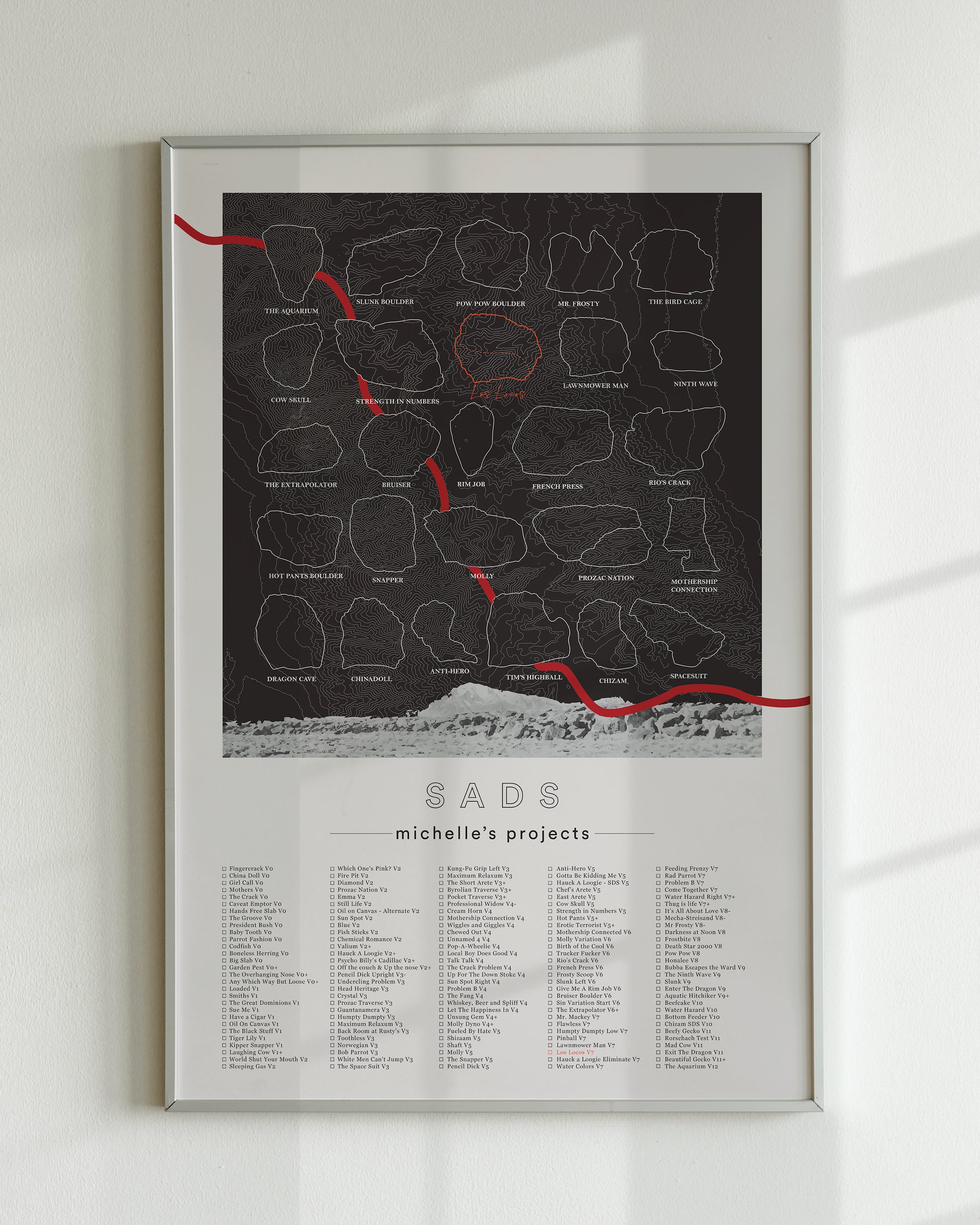A black color, customized rock climbing ticklist poster for the Sads rock climbing location in Bishop, California. The design has an overlay of crag topography, nature landscapes, and traced boulders, the person's name, and a list of bouldering problems at the bottom. Displayed as wall art in a living room.