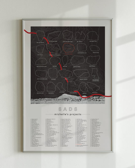 A black color, customized rock climbing ticklist poster for the Sads rock climbing location in Bishop, California. The design has an overlay of crag topography, nature landscapes, and traced boulders, the person's name, and a list of bouldering problems at the bottom. Displayed as wall art in a living room.