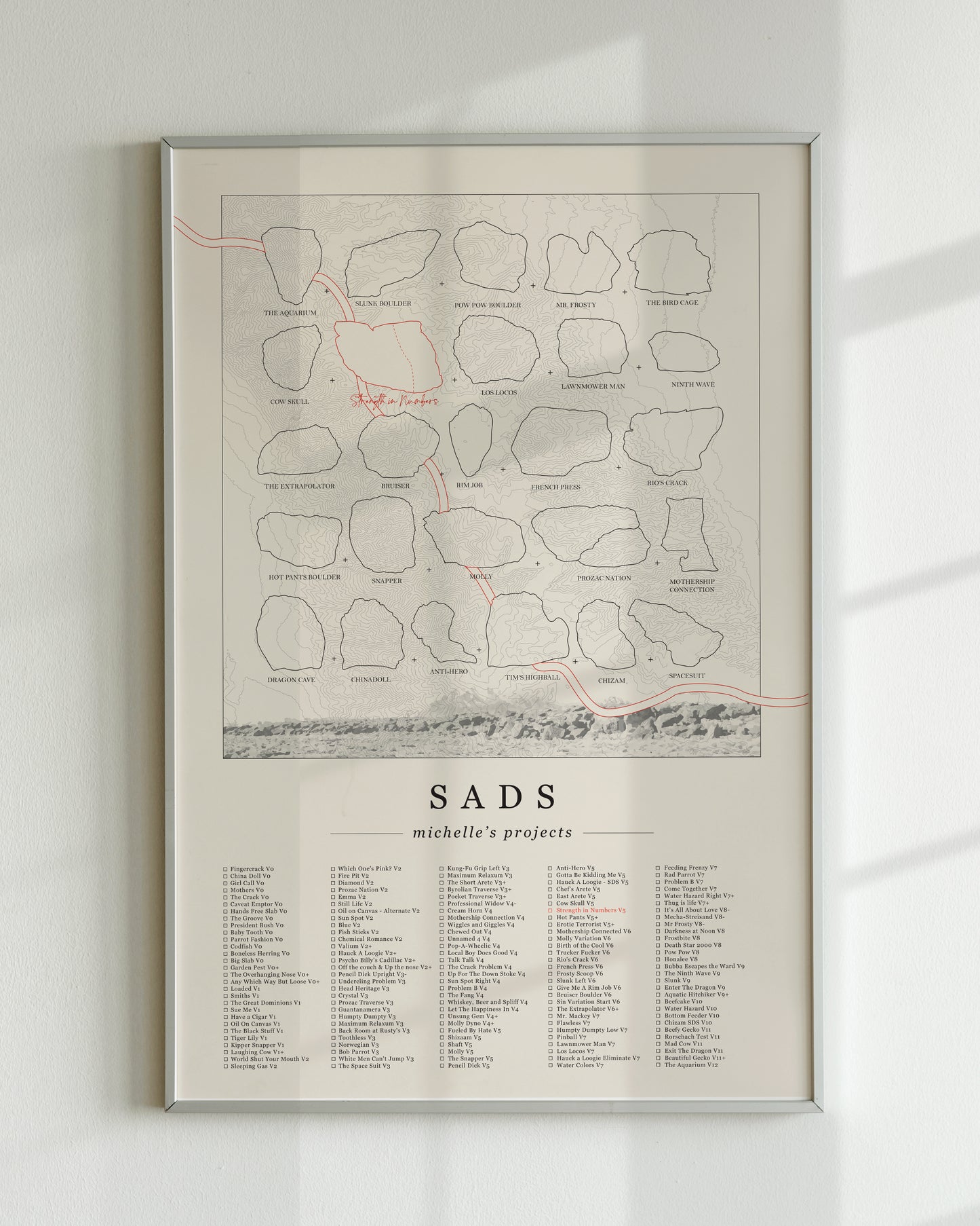 A cream color, customized rock climbing ticklist poster for the Sads rock climbing location in Bishop, California. The design has an overlay of crag topography, nature landscapes, and traced boulders, the person's name, and a list of bouldering problems at the bottom. Displayed as wall art in a living room.
