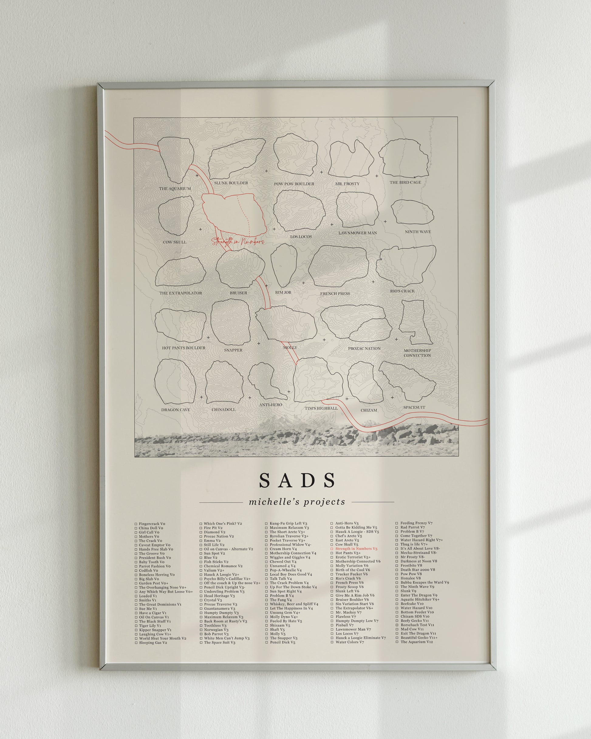 A cream color, customized rock climbing ticklist poster for the Sads rock climbing location in Bishop, California. The design has an overlay of crag topography, nature landscapes, and traced boulders, the person's name, and a list of bouldering problems at the bottom. Displayed as wall art in a living room.